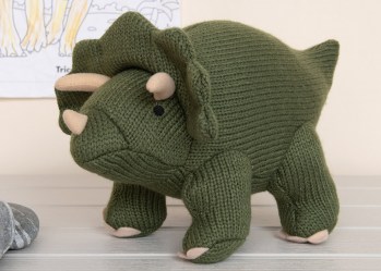 Knitted moss green triceratops dinosaur soft toy on white floorboards with drawing on the wall behind