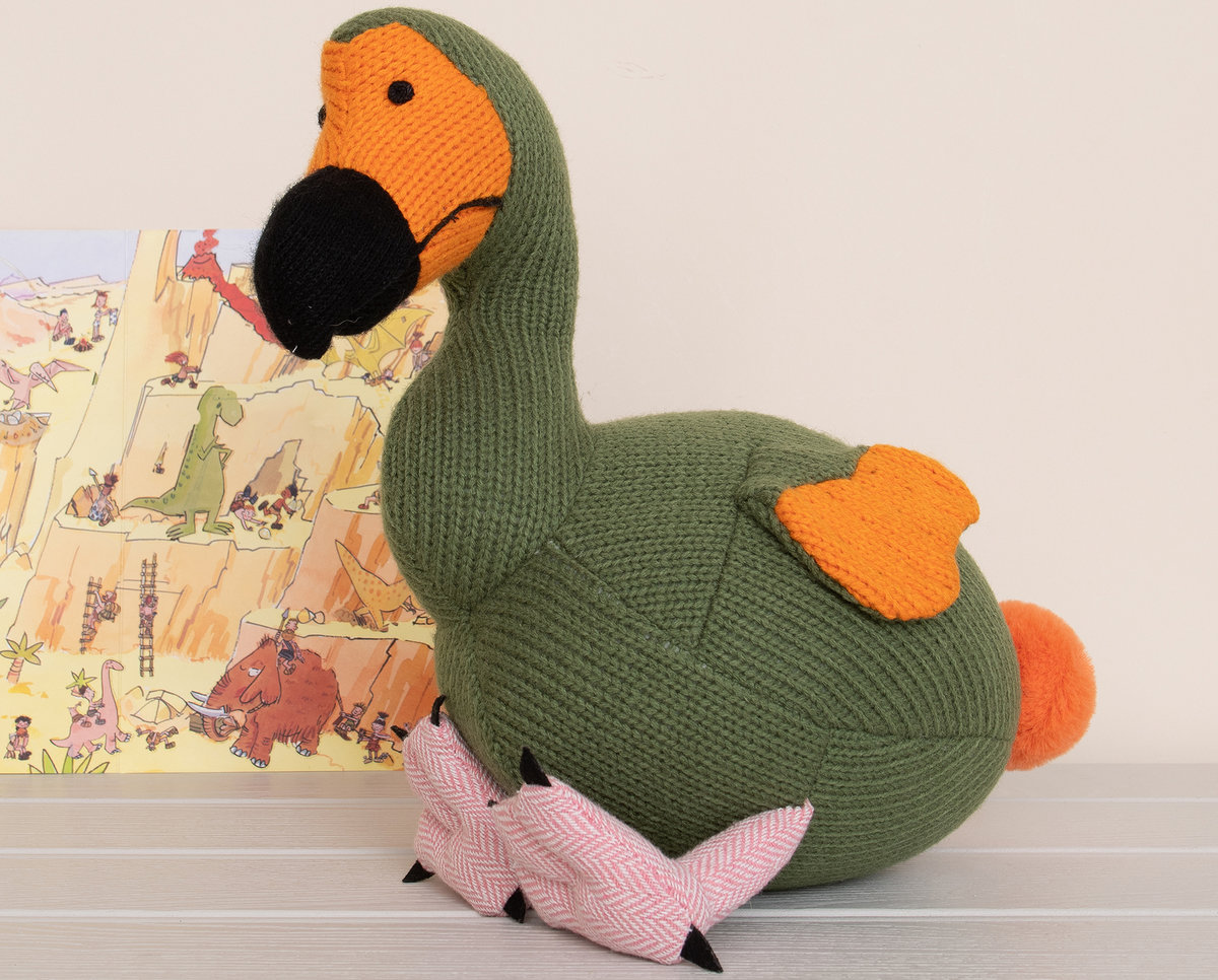 Knitted moss green dodo soft toy with painting of dinosaurs behind